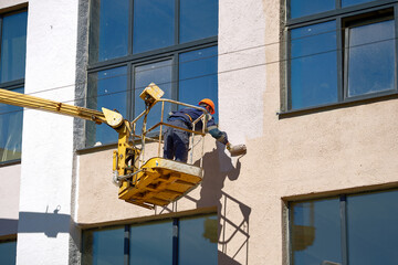 Man paint wall with brush roller in lifting bucket. Painter working on crane platform, paint...