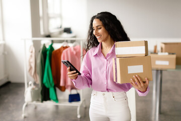 Lady messaging on smartphone holding packed boxes at clothes shop