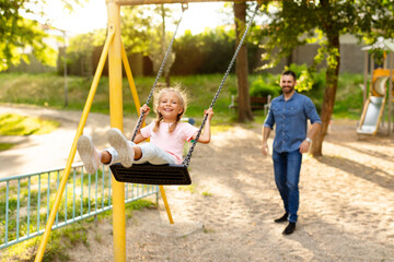 Happy european family of father and daughter spending time at playground, playing and dad pushing child on the swing