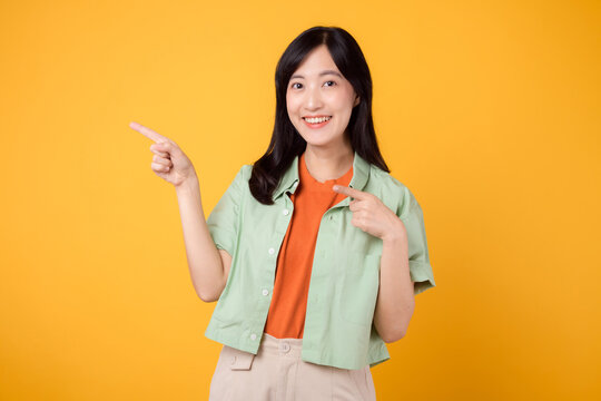 young Asian woman in her 30s wearing a green shirt on an orange shirt, pointing fingers to free copy space. Explore the concept of discount shopping promotion with eye-catching image.