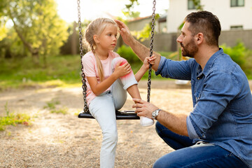 Playground injury. Father talking with daughter sitting on swing, girl touching injured knee and looking at dad - obrazy, fototapety, plakaty
