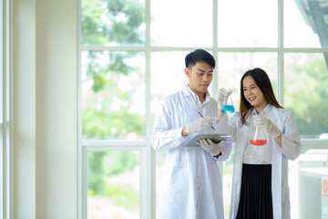 Man and woman scientist standing in laboratory, man holding experiment chart while looking at bottles, woman holding two experiment flasks and looking at them with smile.