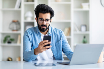 Stressed indian male freelancer looking at smartphone screen in home office