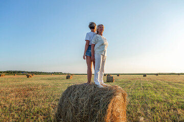 Naklejka na ściany i meble Smiling elderly and young women stand on a roll of hay in a field at sunset. Harvesting, labor and freedom.