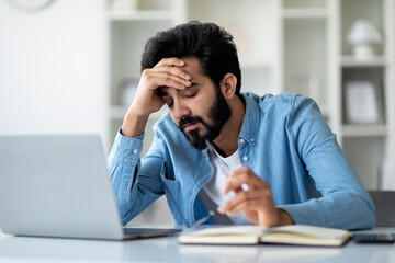 Overworking. Tired Indian Man Sitting At Desk With Laptop And Touching Head