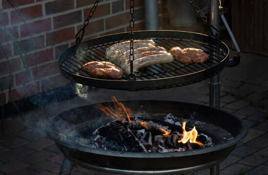 Bratwurst und Nackensteaks auf dem Schwenkgrill auf Holzfeuer vor einer Hauswand mit Regenfallrohr