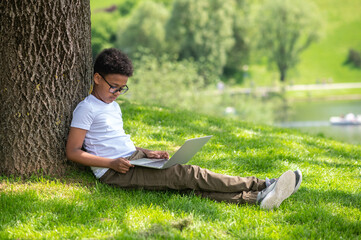 Cute boy sitting in park under tree using laptop to communication with friends or online education.