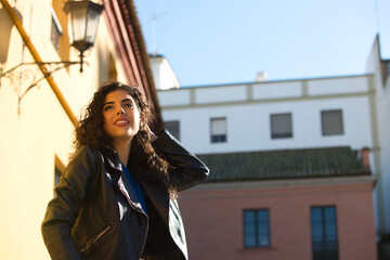 Fototapeta premium Young and beautiful Hispanic brunette woman with curly hair and black leather clothes is sitting on a railing in the street. The woman is happy and smiling.