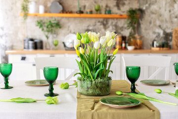 Table festive setting with tulips flowers and stabilization moss in glass vase. Wedding table decoration. Beautiful served table with runner on table, wineglass and plates in living room	