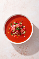 Spanish tomato and watermelon gazpacho cold soup styled and decorated in white plate with ingredients on concrete table in hard light with shadows, top view