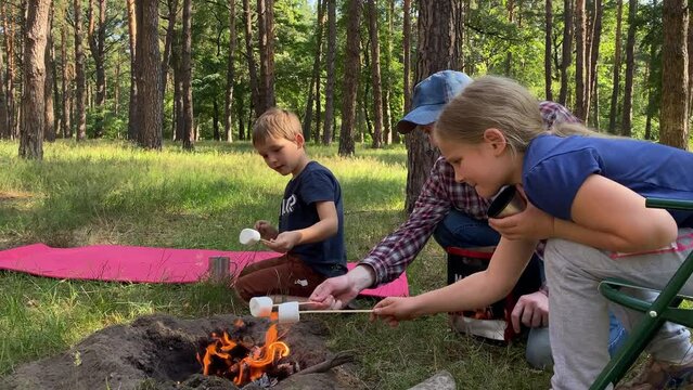 Delightful family camping trip. Father and children gather around campfire to roast marshmallow on wooden sticks in the great outdoors on sunny day. Outdoor cooking while hiking adventure in forest