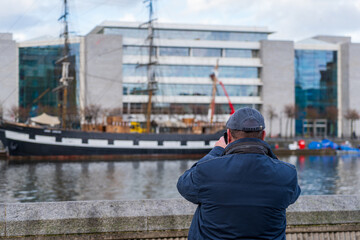 Man wearing a flat cap and blue jacket takes a picture with his smartphone of a sailing ship across...
