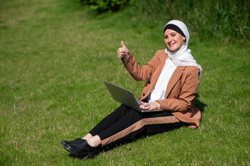 Naklejka na ściany i meble A young woman wearing a hijab sits on a lawn and uses a laptop outdoors and shows a thumbs up.