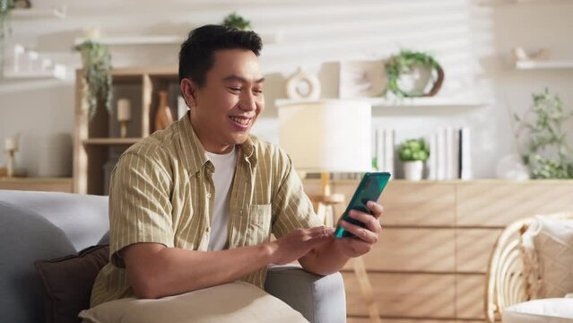 happiness asian adult male man husband smile cheerful spending time using smartphone social media entertain funny content streaming while resting on sofa in living room at home weekend vacation 