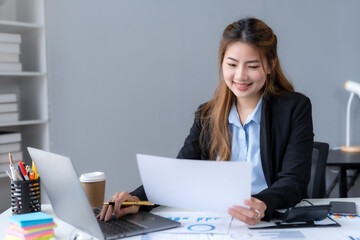 Happy Asian woman accountant at work sitting and taking note of the office report. Asian businesswoman writing a note making a report on investment profits earnings of a company.