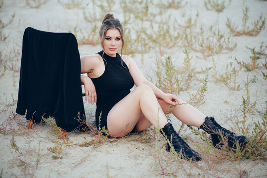 Fashion shot of a plus size model in a black bodysuit sitting on the sand leaning on a chair.