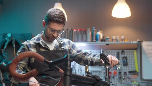 Craftsman repairing bicycle in workshop. Cycling mechanic in glasses and protective apron. Male on background of tools installing saddle on frame. Busy man at workplace in repair shop.