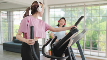 Happy young asian women in sportswear wearing headphones while exercising on the machine at home. Young women talking with mother while training on elliptical machine. Health care concept