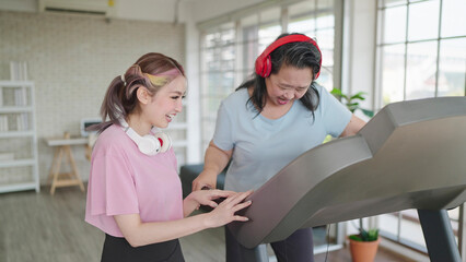 Happy asian elderly women wearing headphones while exercising on treadmill. Young woman helping mother setting elliptical machine for exercising at home. Health care concept