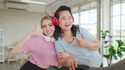 Obraz premium Happy asian elderly women and teenager daughter in sportswear wearing headphones while exercising at home. Smiling and looking at camera. Happy family, Health care concept