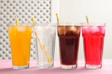 Glasses of different refreshing soda water with ice cubes and straws on pink wooden table