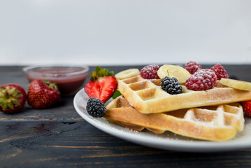 Belgian waffles with summer berries and powdered sugar in a white plate on a dark wooden background.