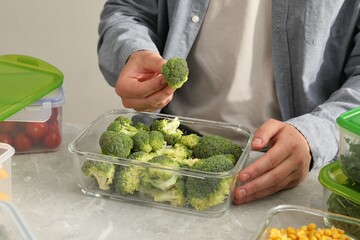 Man putting fresh broccoli into glass container at light grey table, closeup. Food storage