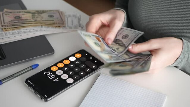 Woman counts dollars and uses phone calculator. Woman calculates expenses of money, manage expenses. Calculation of monthly expenses using phone calculator. Woman saves money for home budget.