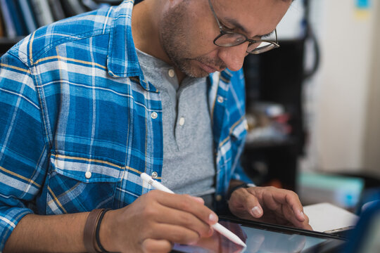 Latin graphic designer working in his office on an illustration tablet, he wears a blue checkered shirt and glasses.