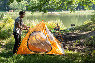 Side view of young bearded man preparing camping tent against river. One shot in series of camping 