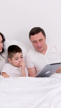 husband wife in bed with little son looking at tablet cartoon interesting game pictures laughing talking communication real people at home love life happy 