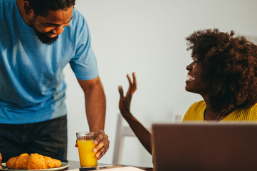 Fototapeta na wymiar An interracial man is surprising his working wife with breakfast while she is taking a break from remote work on a laptop.