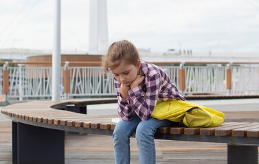 Schoolgirl with sad expression is sitting on bench in park without desire to attend school. An unhappy schoolgirl is sitting in park. Education, lifestyle concept