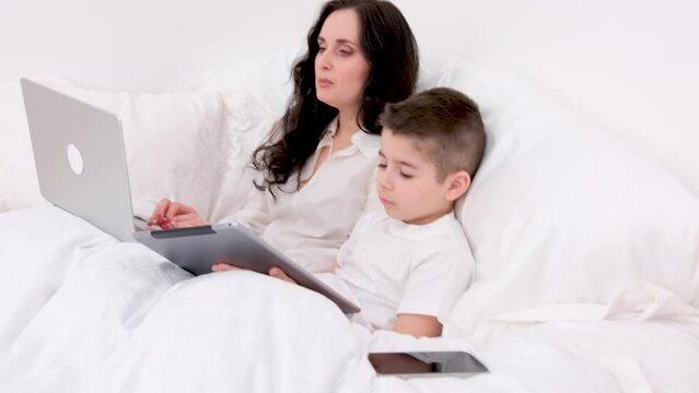 cold lie in bed mother coughs lies next to her son boy of four or five years in the hands of a tablet laptop recover spend time at home work remotely play games be treated