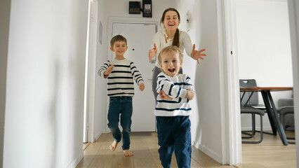 Two little boys and their mother running and laughing on the wooden floor at home. Concept of family love, happiness, and cherished moments spent together.