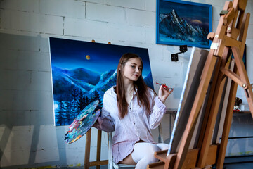 Pretty woman painter in white shirt painting with brush oil paints on canvas in art lab, pensive looking. Portrait of lovely drawing woman artist. Artisanal lifestyle concept. Copy ad text space