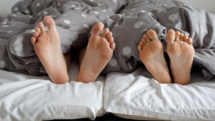 Closeup of couple's feet hiding under the blanket on a cozy bed. Concept of romantic evening,...