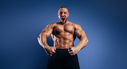 Fototapeta na wymiar Emotional bodybuilder screaming and showing muscles on blue background. Fit man with muscular body