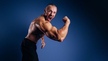 Emotional bodybuilder screaming and showing muscles on blue background. Fit man with muscular body