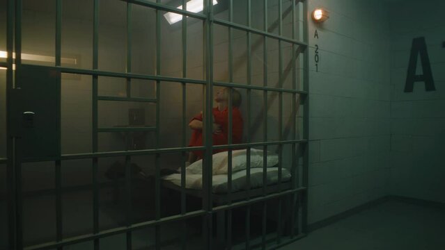 Dolly shot of female prisoner in orange uniform sitting on the bed in jail cell, looking at barred window. Woman serves imprisonment term for crime in prison. Depressed criminal in detention center.