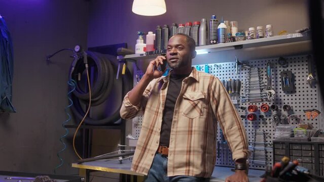 African American Craftsman Talking By Phone. Repair Man Leaning On Table At Background Of Rack With Tools. Cycling Mechanic Standing At Workshop At Background Of Repair Equipment. Ethnic Diveristy.