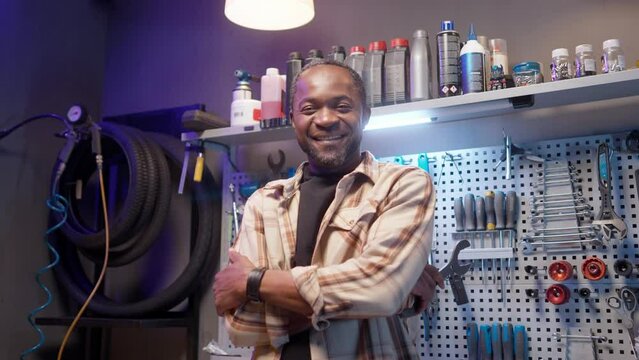 African American caftsman smiling with teeth. Repair man posing at background of tools. Cycling mechanic standing at workshop and looking at camera. Satisfied male with crossed arms at repair shop.