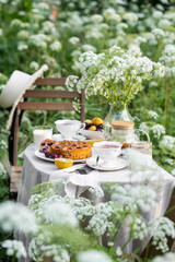Summer elegant picnic. Woman in white dress purring tea from teapot at wooden table in garden. Beautiful dishwater, white porcelain, tablecloth, homemade cherry pie.