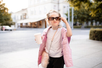 Fototapeta premium Cute kid girl 4-5 year old wear sun glasses and trendy casual clothes holding coffee cup having fun at city street over sunset outdoor. Portrait of cheerful and happy child. Summer season.