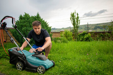 A man in blue shorts and a black T-shirt inspects the condition of a lawnmower in blue and black colors against a background of trees and a fence