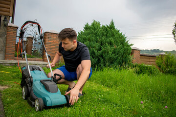 A man in blue shorts and a black T-shirt inspects the condition of a lawnmower in blue and black colors against a background of trees and a fence