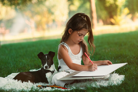 A beautiful, sweet little girl is spending time with her pet dog and drawing a picture with colored pencils in the park