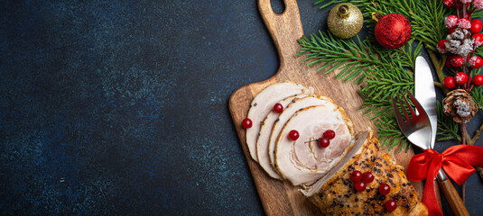 Christmas baked ham sliced with red berries and festive decorations on wooden cutting board, dark rustic background from above. Christmas and New Year holiday dinner with baked pork, space for text - 625333655