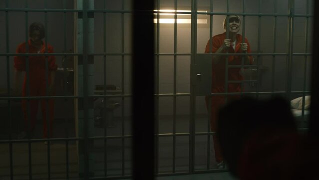 One aggressive female prisoner in orange uniform yells at inmate and jumps on bars, another nervously walks in prison cell. Women serve imprisonment terms for crimes in jail. Correctional facility.