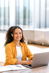 Vertical photo of a successful curly haired girl, brazilian or hispanic nationality, outsourcing employee or student, uses laptop to work or study online while sits outdoors, looks at camera, smiles